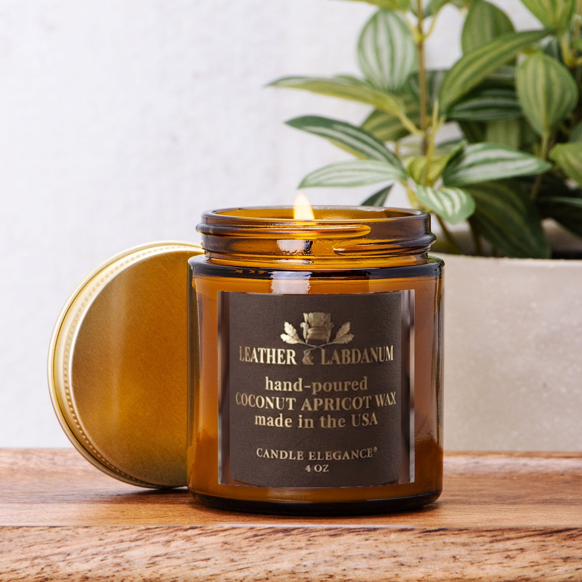 An amber glass jar 'Leather & Labdanum' candle with its lid off, showing a lit flame. The candle is placed on a wooden surface with a potted green plant in the background.