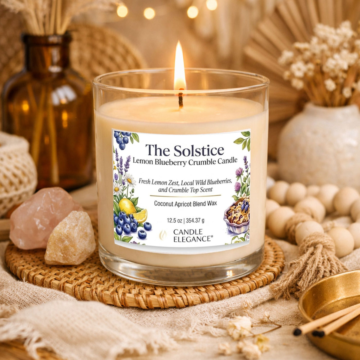 A lit candle in a clear glass jar sits on a woven coaster surrounded by dried flowers, crystals, and warm decorative props. The label reads 'The Solstice Lemon Blueberry Crumble Candle' by Candle Elegance.