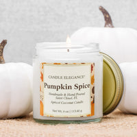 A lit Candle Elegance 'Pumpkin Spice' scented candle in a clear glass jar, shown with its gold metal lid leaning against it. The jar is styled on a burlap surface with white pumpkins in the background.