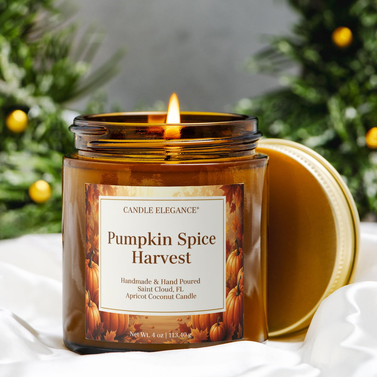 A lit amber glass jar candle labeled 'Pumpkin Spice Harvest' by Candle Elegance, with the gold lid resting beside it on white fabric. The background features soft-focus green foliage with yellow ornaments.