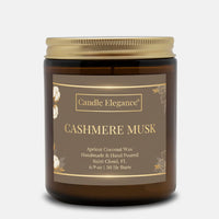 A Candle Elegance Cashmere Musk scented candle in an amber glass jar with a gold metal lid and a brown label, shown on a light gray background.