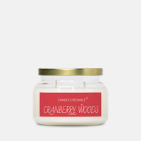 A jar candle with white wax, a gold metal lid, and a red label reading 'CRANBERRY WOODS' on a light gray background.