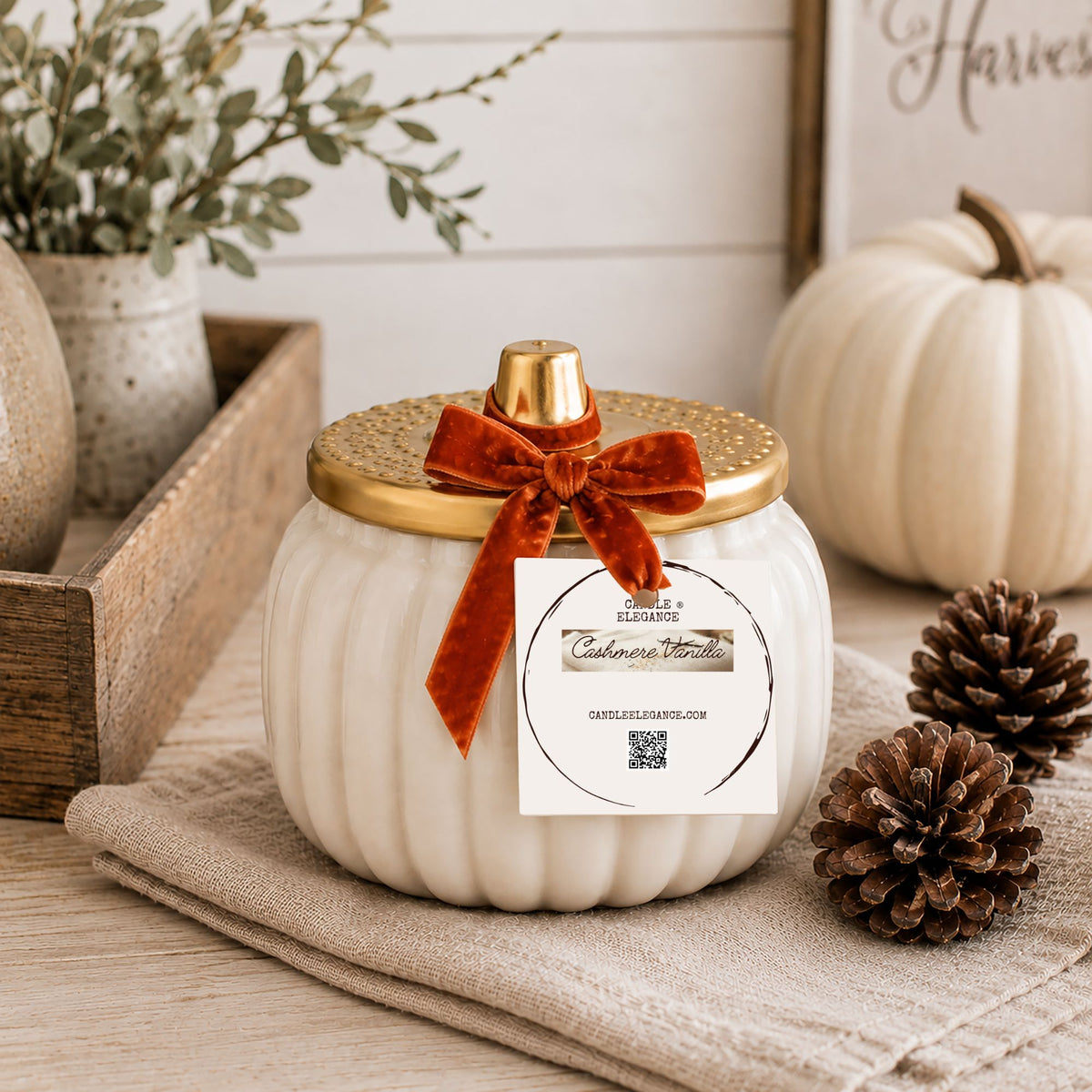 A ribbed white decorative ceramic pumpkin jar candle by Candle Elegance, with a gold lid and a rust-orange velvet bow, styled on a beige cloth with pumpkins, pinecones, and a wooden tray in a fall-themed setting.