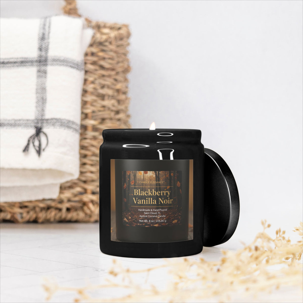 A black jar candle with a lit wick and a hinged side lid, labeled 'Blackberry Vanilla Noir' by Candle & Co, styled on a white surface with a woven basket and white towel in the background.