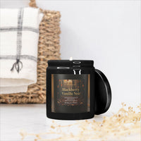 A black jar candle with a lit wick and a hinged side lid, labeled 'Blackberry Vanilla Noir' by Candle & Co, styled on a white surface with a woven basket and white towel in the background.
