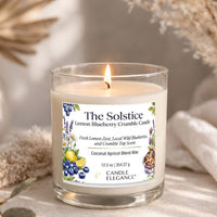 A lit candle in a clear glass jar with a white label reading 'The Solstice Lemon Blueberry Crumble Candle' and 'Candle Elegance' at the bottom. The candle is photographed on a soft neutral surface with dried floral props blurred in the background.