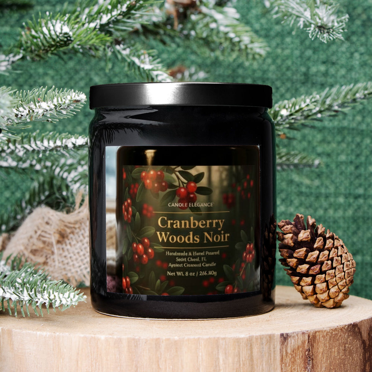 A black glass jar candle labeled 'Candle Elegance Cranberry Woods Noir' sits on a wooden surface with evergreen branches, burlap, and a pinecone around it against a green textured backdrop.