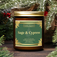 A jar candle labeled 'Sage & Cypress' by Candle Eleganee, set on a wooden surface surrounded by pine branches and red berries. The candle jar is amber glass with a gold lid and a dark green label with gold leaf accents.