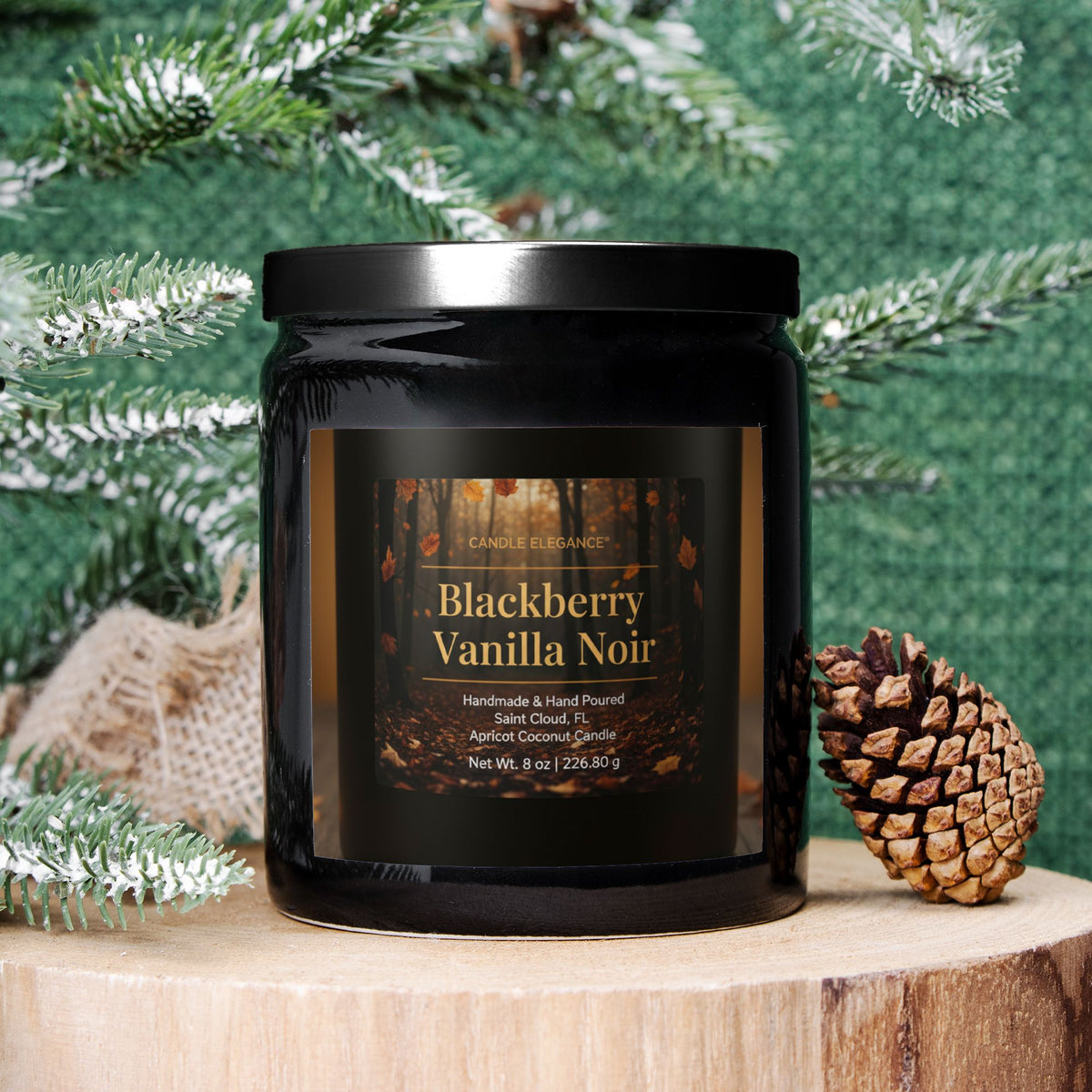 A black glass jar candle labeled 'Blackberry Vanilla Noir' by Candle & Equilibrium, styled on a wooden slice with a pinecone and frosted green pine branches against a textured green backdrop.