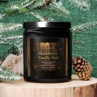 A black glass jar candle labeled 'Blackberry Vanilla Noir' by Candle & Equilibrium, styled on a wooden slice with a pinecone and frosted green pine branches against a textured green backdrop.