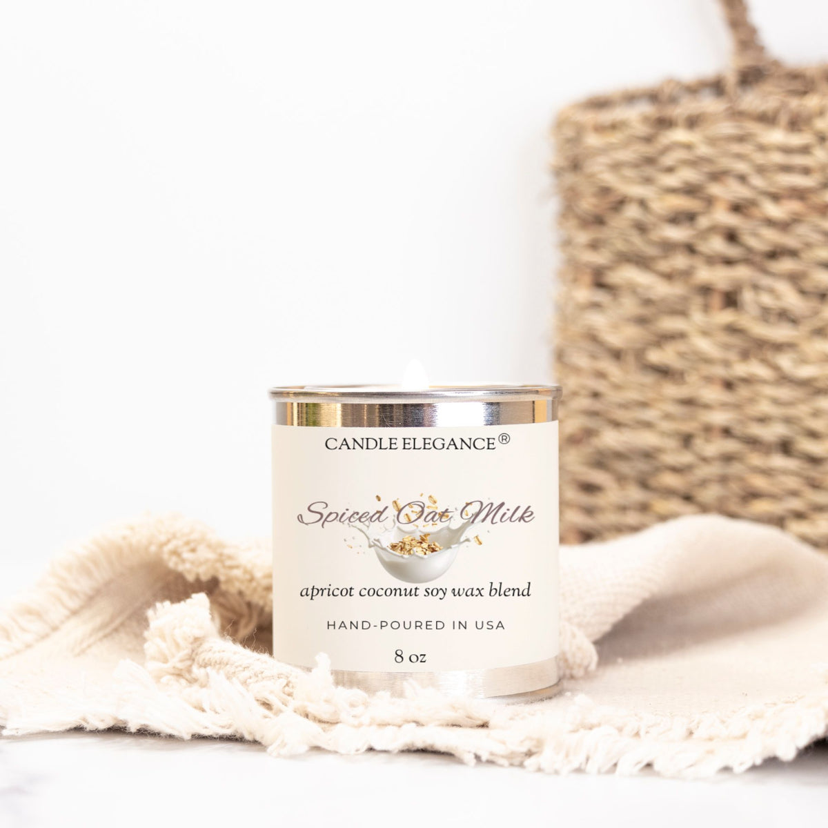 An 8 oz candle by Candle Elegance in a white jar with a silver lid, styled on a cream fringed cloth with a woven basket in the background.