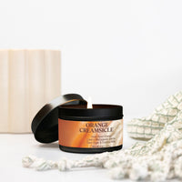 An open black candle tin with a lit wick and a warm orange label reading 'ORANGE CREAMSICLE,' styled on a white surface with a cream ribbed container in the background and soft textured props around it.