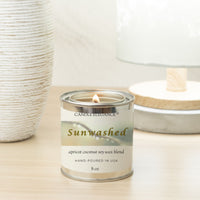 A lit 8 oz candle in a silver metal tin with a cream and green label reading 'CandleLounge Sunwashed apricot coconut soy wax blend'. The candle sits on a light wood surface with a textured white vase on the left and a soft-focus lamp in the background.