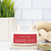 A double-wick candle in a clear glass jar with white wax and a red label reading 'Cranberry Woods' and 'Sindige Egecusis', styled on a marble coaster with a potted plant and wooden decor in the background.
