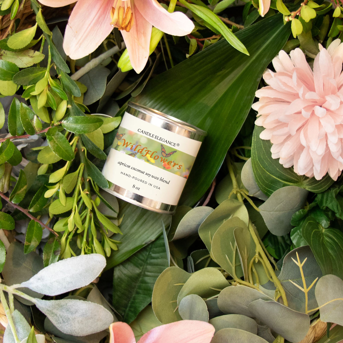A candle labeled 'Wildflowers' by Candleflavourz, nestled among green leaves and pink flowers in a styled flat-lay arrangement.
