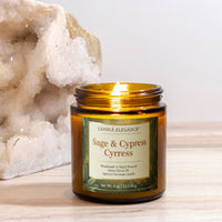 An amber glass jar candle labeled 'Candle Elegance Sage & Cypress' sits on a light wood surface with a large white crystal geode prop in the background.