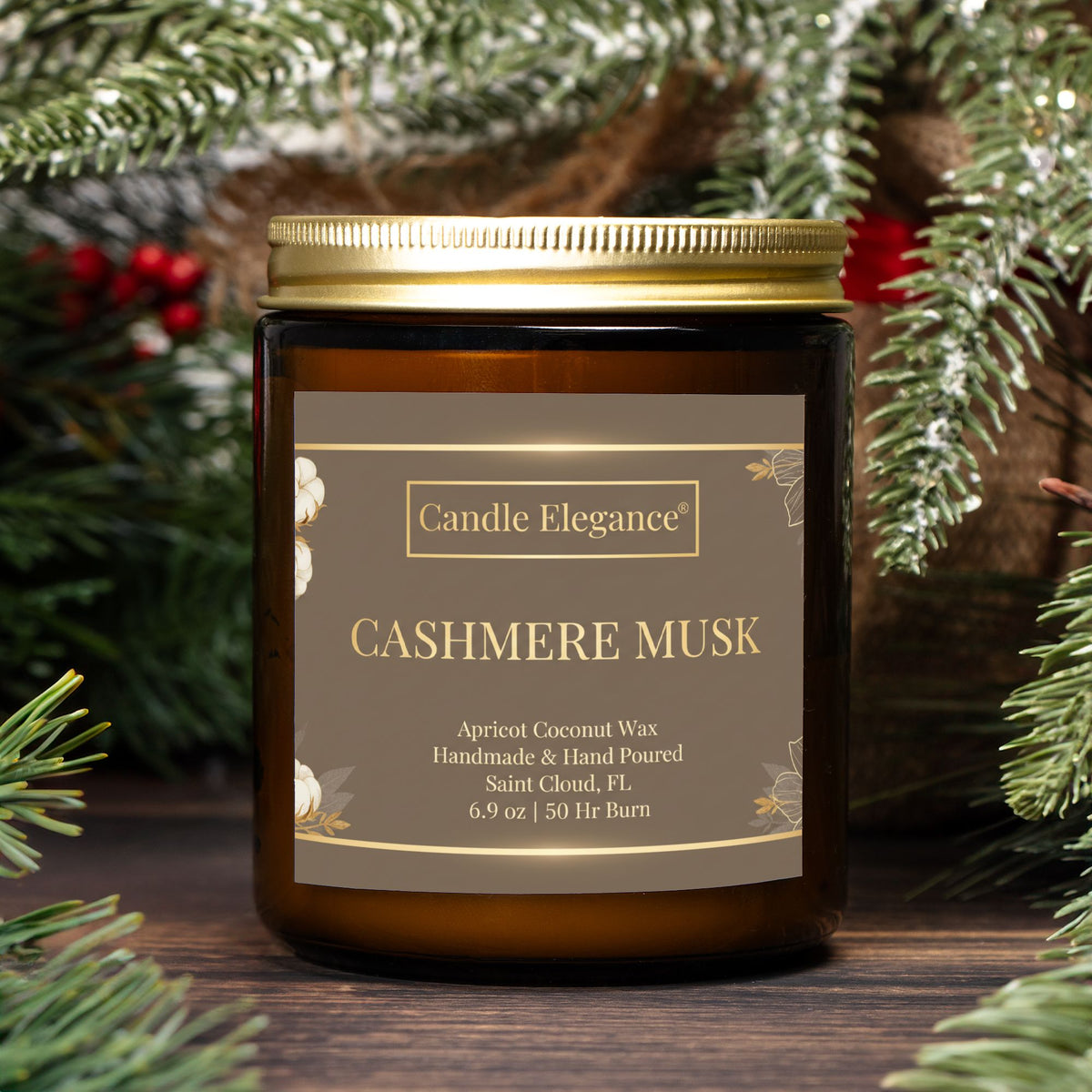 An amber glass jar candle with a gold metal lid and a brown label reading 'Candle Elegance Cashmere Musk,' styled with pine branches and red berries on a wooden surface.