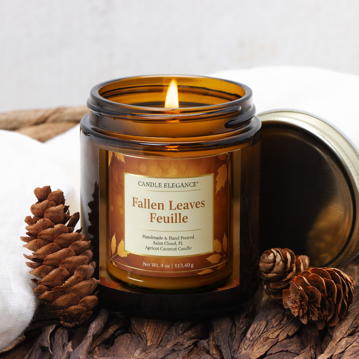 A lit amber glass jar candle labeled Candle Elegance Fallen Leaves Feuille, styled on a rustic wood surface with pinecones and a white cloth, with the metal lid leaning behind it.