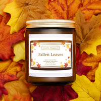 A jar candle labeled 'Fallen Leaves' by Candle Elegance, placed on a bed of colorful autumn leaves. The candle jar is amber brown with a gold lid and a white label featuring red and orange maple leaves.