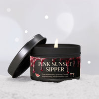 A black metal candle tin with its lid leaning against it, showing a lit candle inside. The label reads 'PINK SUNSET SIPPER' with watermelon and citrus graphics, set on a white textured surface against a soft gray bokeh background.