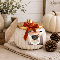 A white ribbed pumpkin-shaped jar candle with a gold lid and a rust-orange velvet bow, displayed on a beige cloth with pinecones and a small white pumpkin in a rustic fall setting.
