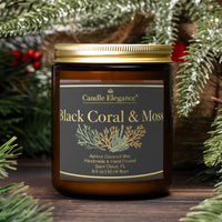 An amber glass jar candle with a gold lid and a dark gray label reading 'Candle Elegance Black Coral & Moss' is centered on a wooden surface, surrounded by pine branches and red berries.