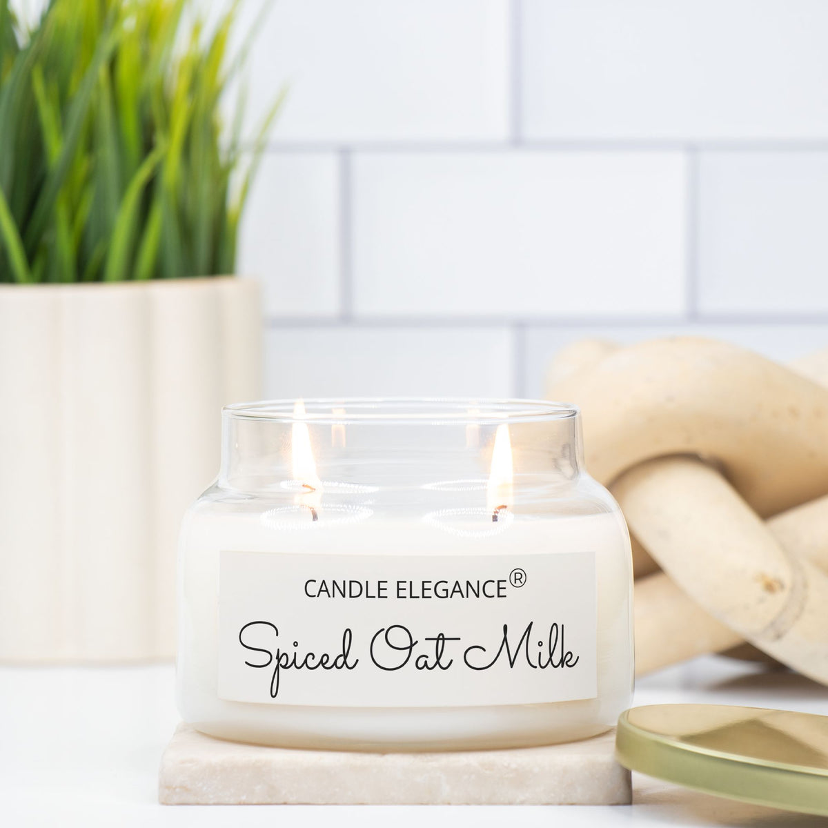 A lit double-wick candle in a clear glass jar with a white label reading 'CANDLE ELEGANCE Spiced Oat Milk,' styled on a marble coaster with a potted plant and woven decor in the background.