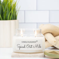 A lit double-wick candle in a clear glass jar with a white label reading 'CANDLE ELEGANCE Spiced Oat Milk,' styled on a marble coaster with a potted plant and woven decor in the background.