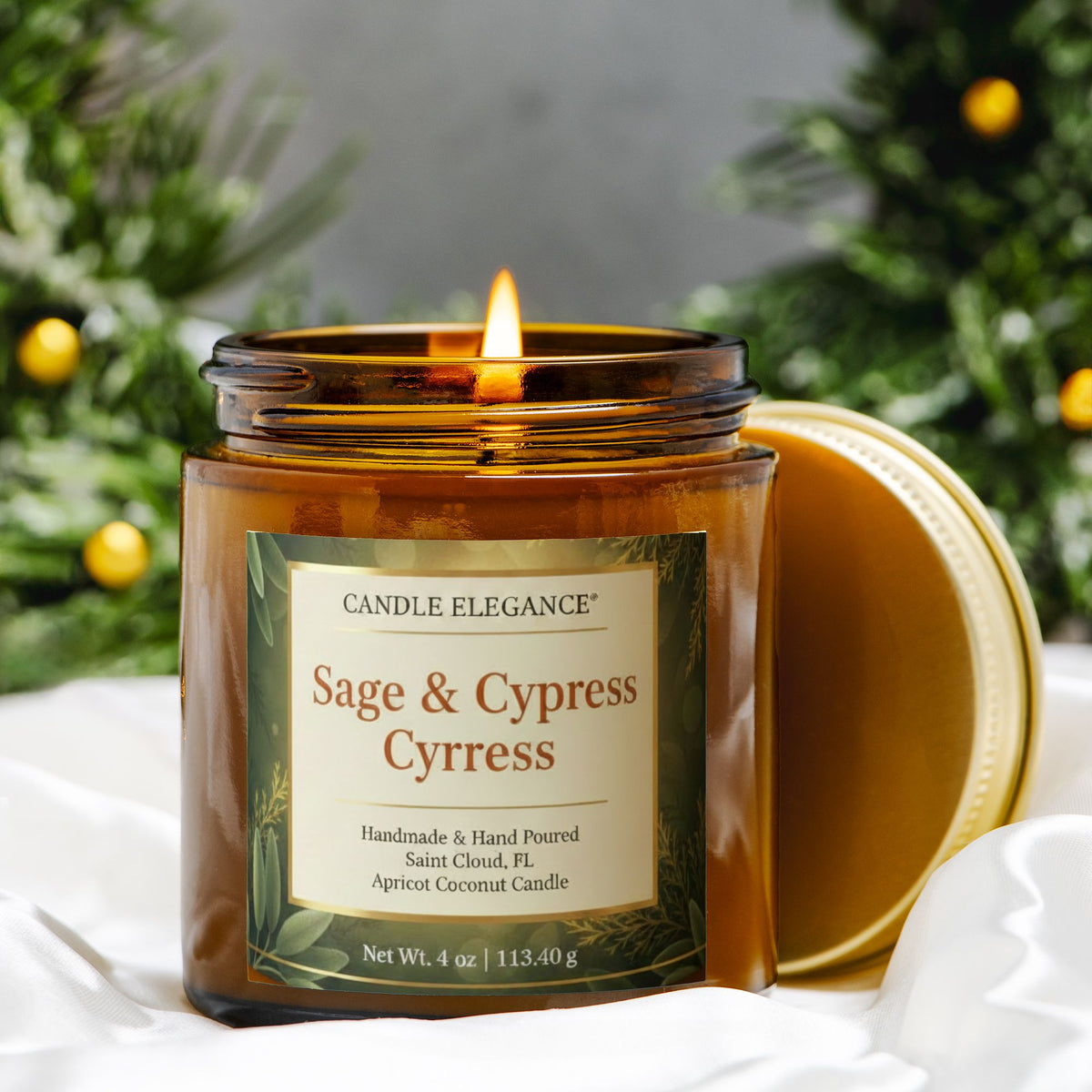 A lit amber glass jar candle with the lid resting beside it on white fabric, set against a blurred holiday greenery background. The front label reads Candle Elegance Sage & Cypress Cypress.