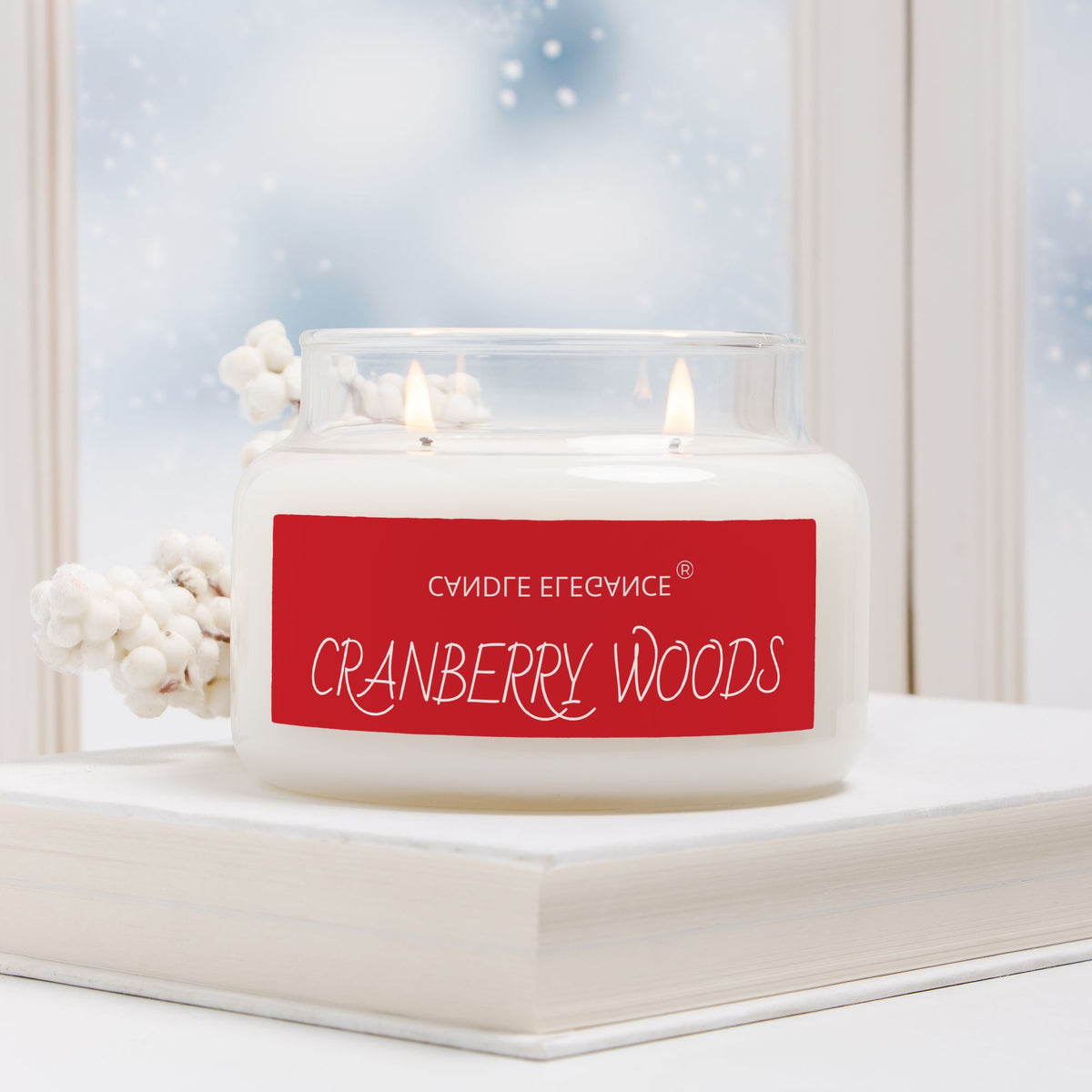 A white wax candle in a clear glass jar sits on a white book, with two lit flames visible. The front label is bright red with white text reading 'Cranberry Woods' and additional Cyrillic text above it.