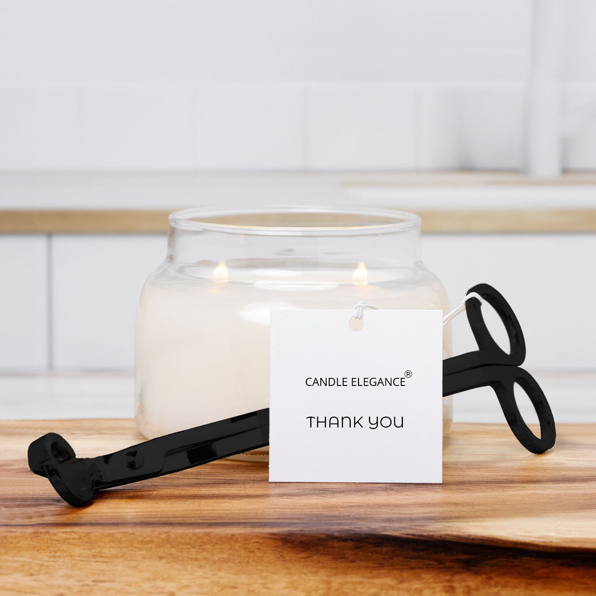 A lit candle in a clear glass jar with a black scissor-style candle holder resting in front on a wooden surface. A white tag reading 'CANDLE ELEGANCE THANK YOU' is attached, and the background shows a softly blurred white tiled interior.