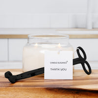 A lit candle in a clear glass jar with a black scissor-style candle holder resting in front on a wooden surface. A white tag reading 'CANDLE ELEGANCE THANK YOU' is attached, and the background shows a softly blurred white tiled interior.