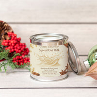 A small metal candle tin labeled Candle Elegance Spiced Oat Milk sits on a whitewashed wood surface with a lid leaning beside it. Red berries, greenery, and a pinecone are visible on the left, and part of a green ornament appears on the right, creating a styled holiday scene.