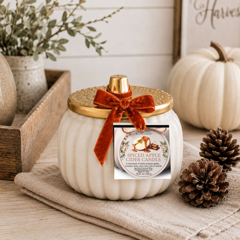 A ribbed white ceramic candle jar shaped like a pumpkin, topped with a gold lid and a rust-brown velvet bow. A product label for 'Dishlight' 'Spiced Apple Cider Candle' is attached to the front.