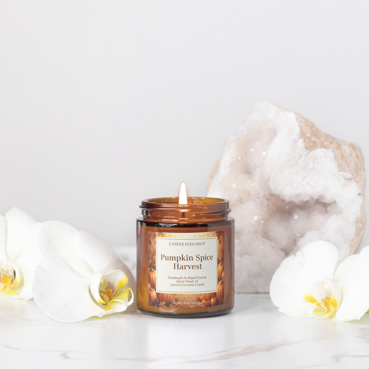 An amber glass jar candle labeled 'Pumpkin Spice Harvest' by Candle Elegance, styled on a white marble surface with white orchids and a large geode crystal in the background.