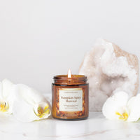 An amber glass jar candle labeled 'Pumpkin Spice Harvest' by Candle Elegance, styled on a white marble surface with white orchids and a large geode crystal in the background.