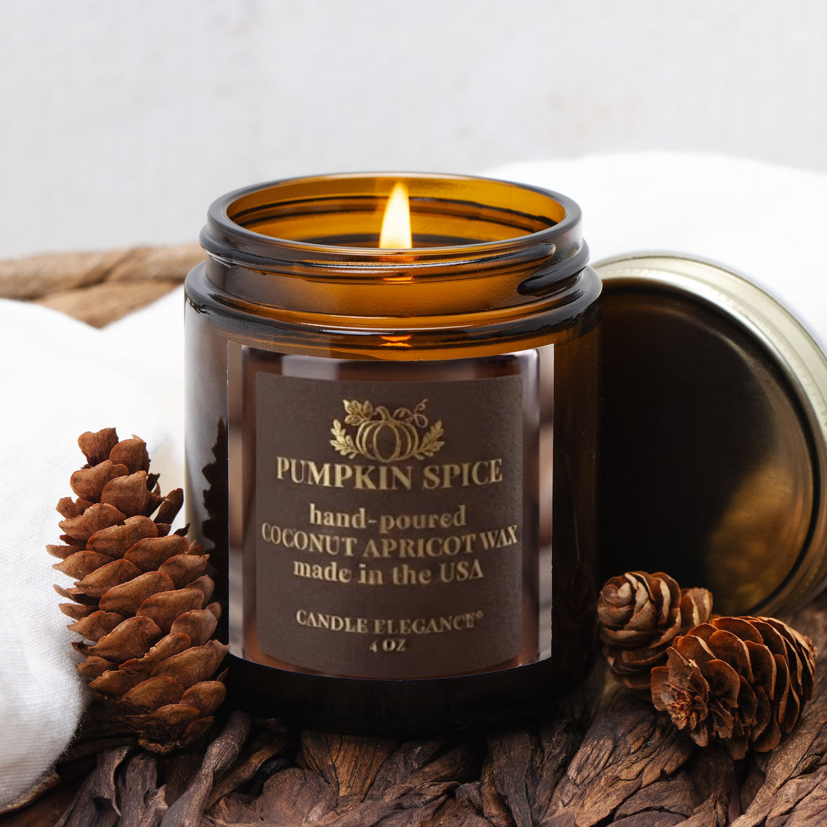 A lit amber glass candle jar labeled 'Pumpkin Spice' by Candle Elegance, shown with its metal lid beside it on a rustic surface with pinecones and a white cloth.