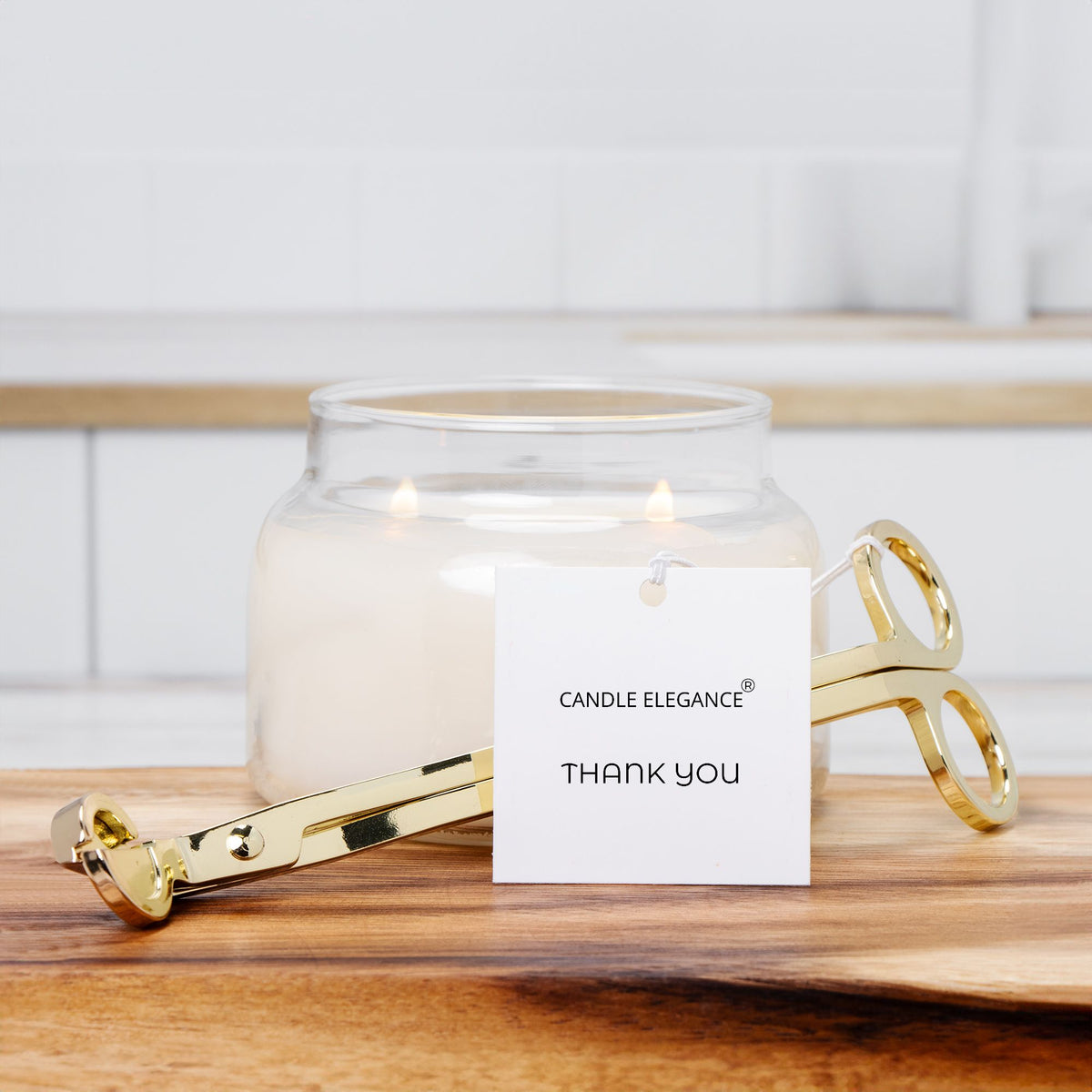 A gold metal candle tongs tool resting in front of a lit white candle in a clear glass jar, with a white tag reading 'CANDLE ELEGANCE THANK YOU' on a wooden surface against a bright tiled background.