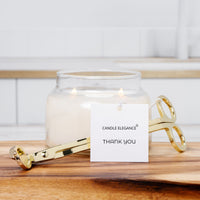 A gold metal candle tongs tool resting in front of a lit white candle in a clear glass jar, with a white tag reading 'CANDLE ELEGANCE THANK YOU' on a wooden surface against a bright tiled background.