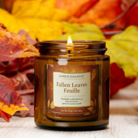 A lit amber glass jar candle labeled 'Candle Elegance Fallen Feuille' sits on a light wood surface surrounded by colorful autumn leaves.