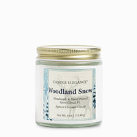 A 4 oz glass jar candle with a gold metal lid and a white label reading 'Woodland Snow' by Candle Elegance, shown on a clean white background.
