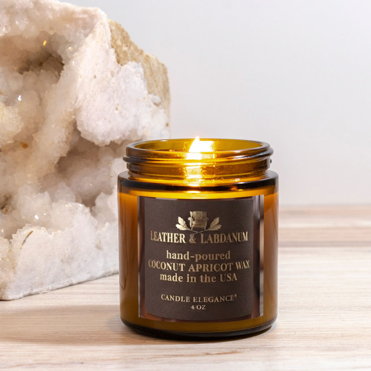 An amber glass jar candle with a lit flame, featuring a dark brown label that reads 'LEATHER & LABDANUM hand-poured COCONUT APRICOT WAX made in the USA'. The candle is set on a light wood surface next to a large white geode crystal, with a soft neutral background.