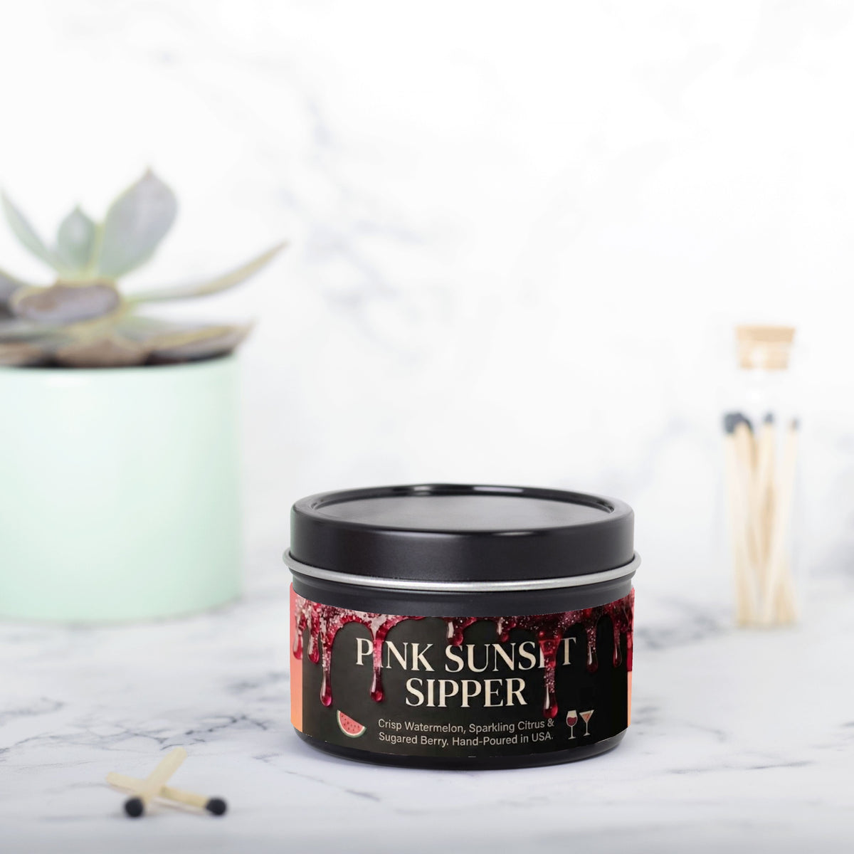 A black metal tin of 'Pink Sunset Sipper' gummy candies sits on a white marble surface with a blurred succulent and a glass of straws in the background.