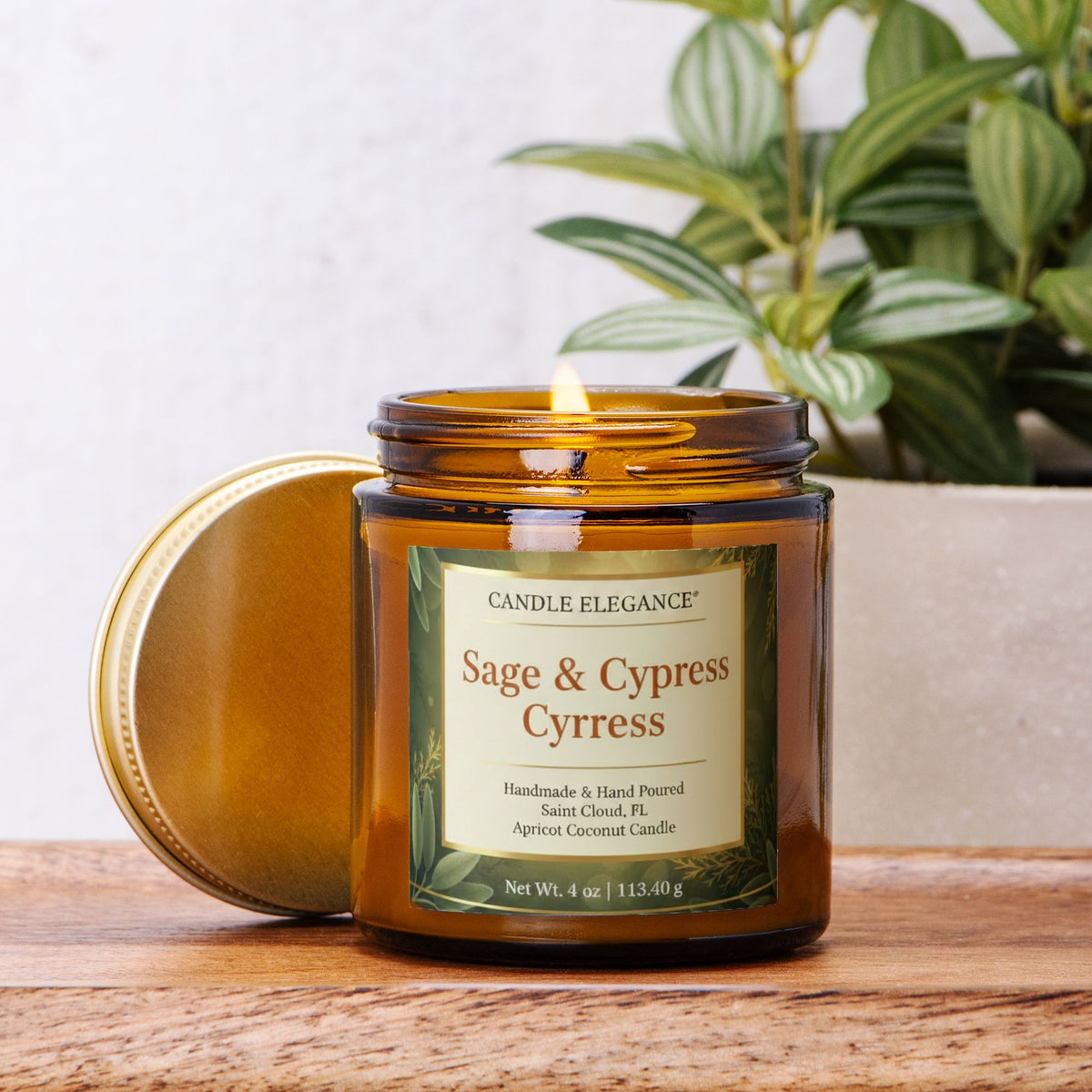 An amber glass jar candle with its lid leaning beside it on a wooden surface. The front label reads Candle Elegance Sage & Cypress Cypress, and a lit flame is visible inside the jar.