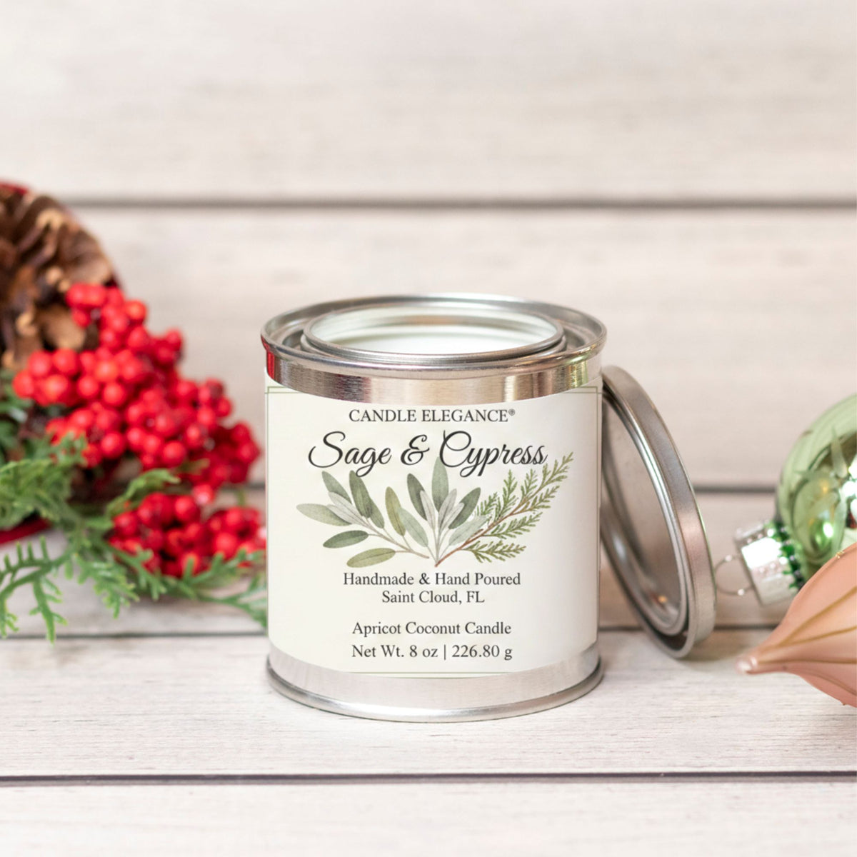 An 8 oz metal tin candle labeled 'Candle Elegance Wild Flowers' sits on a light wood surface with its lid leaning beside it. The scene is styled with red berries, greenery, and a pinecone on a whitewashed wood background.