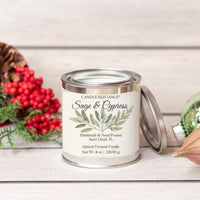 An 8 oz metal tin candle labeled 'Candle Elegance Wild Flowers' sits on a light wood surface with its lid leaning beside it. The scene is styled with red berries, greenery, and a pinecone on a whitewashed wood background.