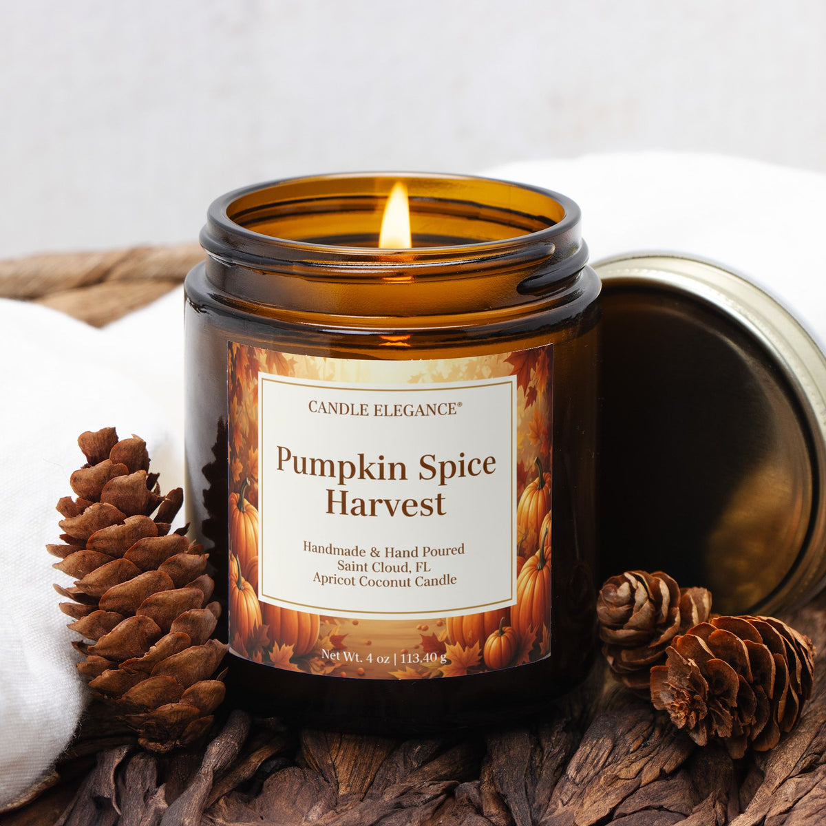 A lit amber glass jar candle labeled Candle Elegance Pumpkin Spice Harvest, styled with pinecones and dried leaves on a rustic surface with a white cloth in the background.
