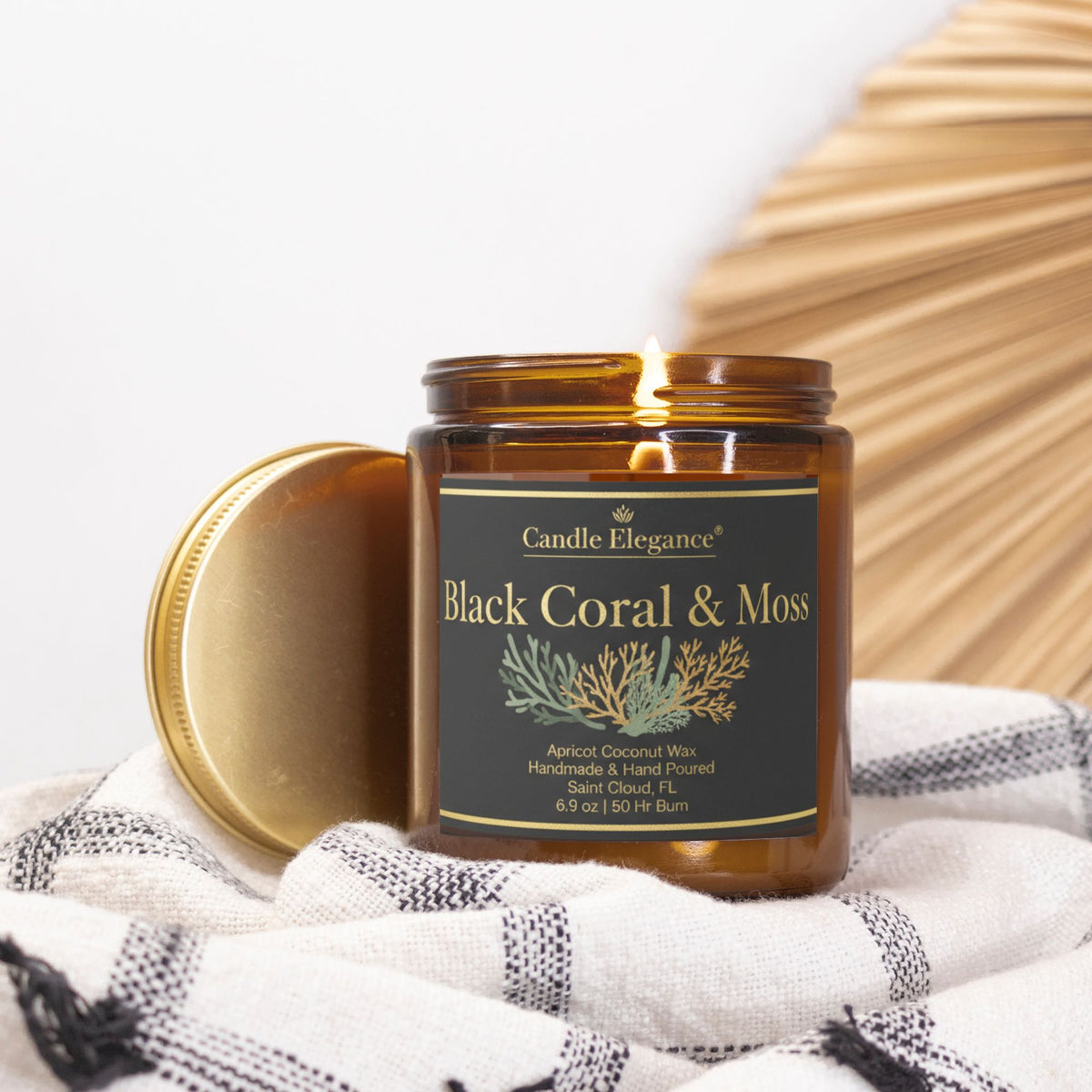 An amber glass jar candle with its gold lid leaning beside it, placed on a white and gray checkered cloth. The candle jar has a black label with gold lettering that reads 'Black Coral & Moss' and 'Candle Elegance'.