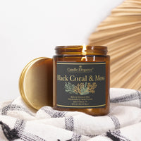 An amber glass jar candle with its gold lid leaning beside it, placed on a white and gray checkered cloth. The candle jar has a black label with gold lettering that reads 'Black Coral & Moss' and 'Candle Elegance'.