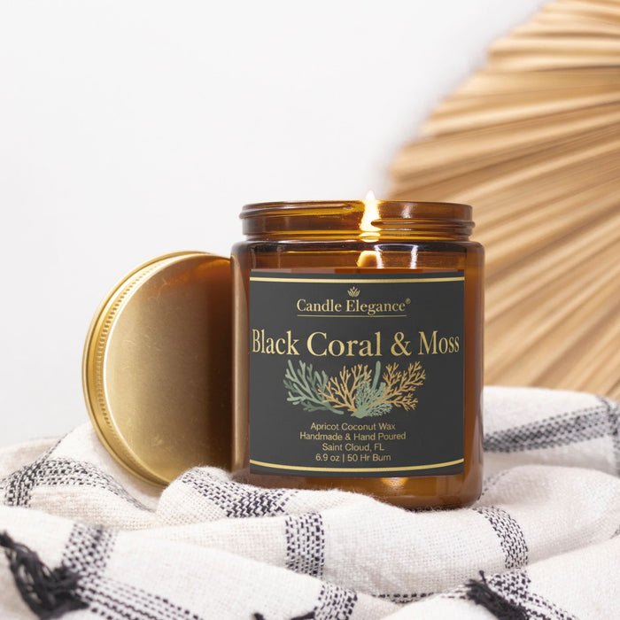 An amber glass jar candle with its gold lid leaning beside it, placed on a white and gray checkered cloth. The candle jar has a black label with gold lettering that reads 'Black Coral & Moss' and 'Candle Elegance'.